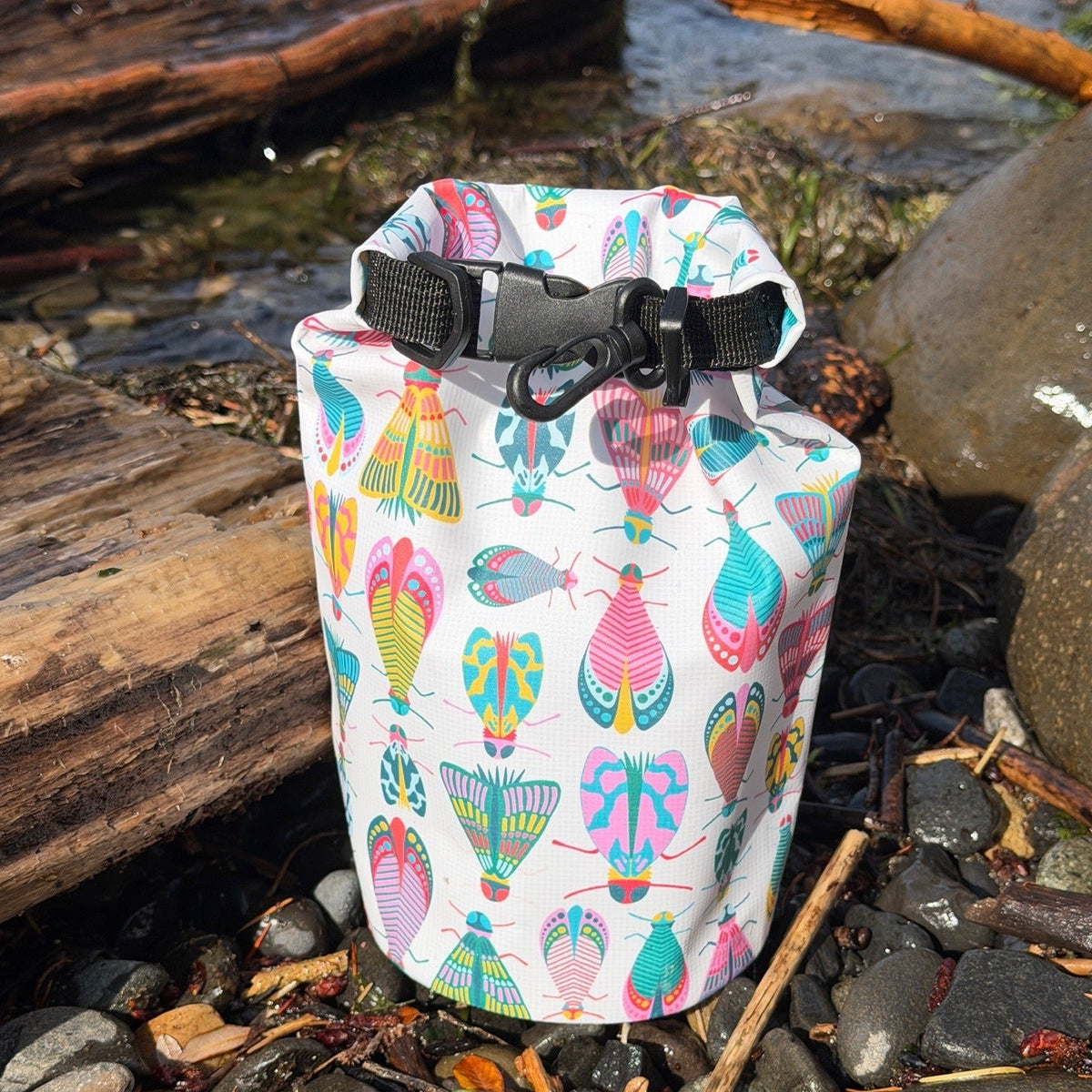 A roll-top dry bag with a colorful pattern of stylized moths and fish on a white background, standing on a rocky shoreline with logs and water in the background.