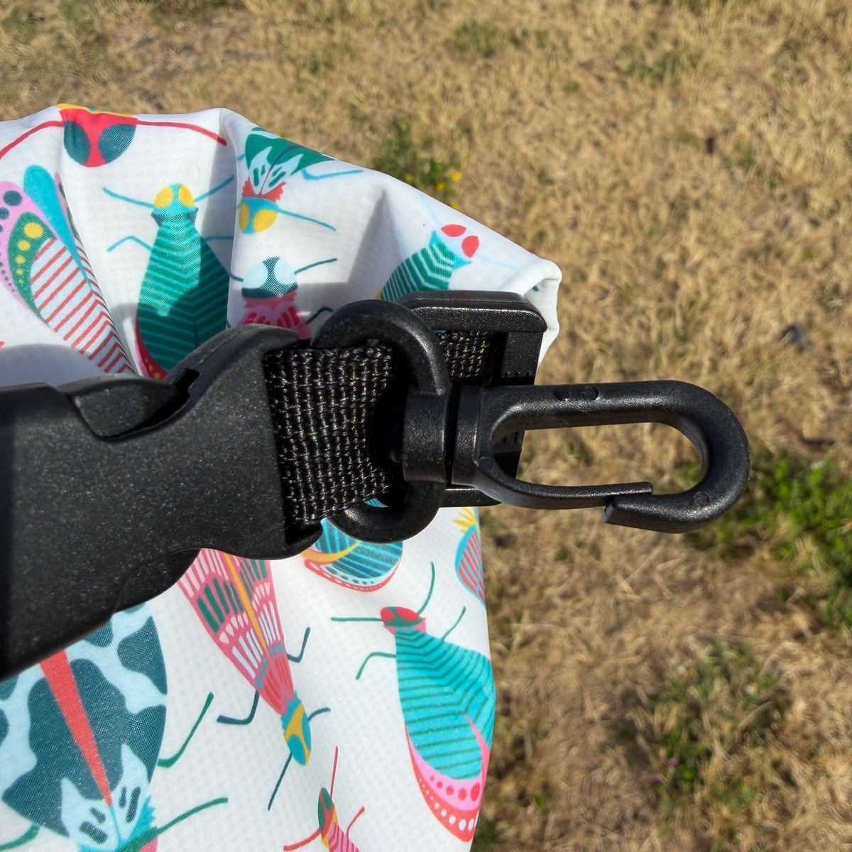 Close-up of a black metal carabiner clipped to a white waterproof bag printed with colorful insect patterns, set against a dry grassy outdoor background.