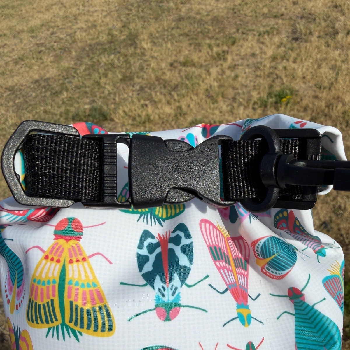 A close-up of a roll-top bag with a colorful insect pattern on a white background, featuring a black plastic buckle closure, set against a dry grassy outdoor background.