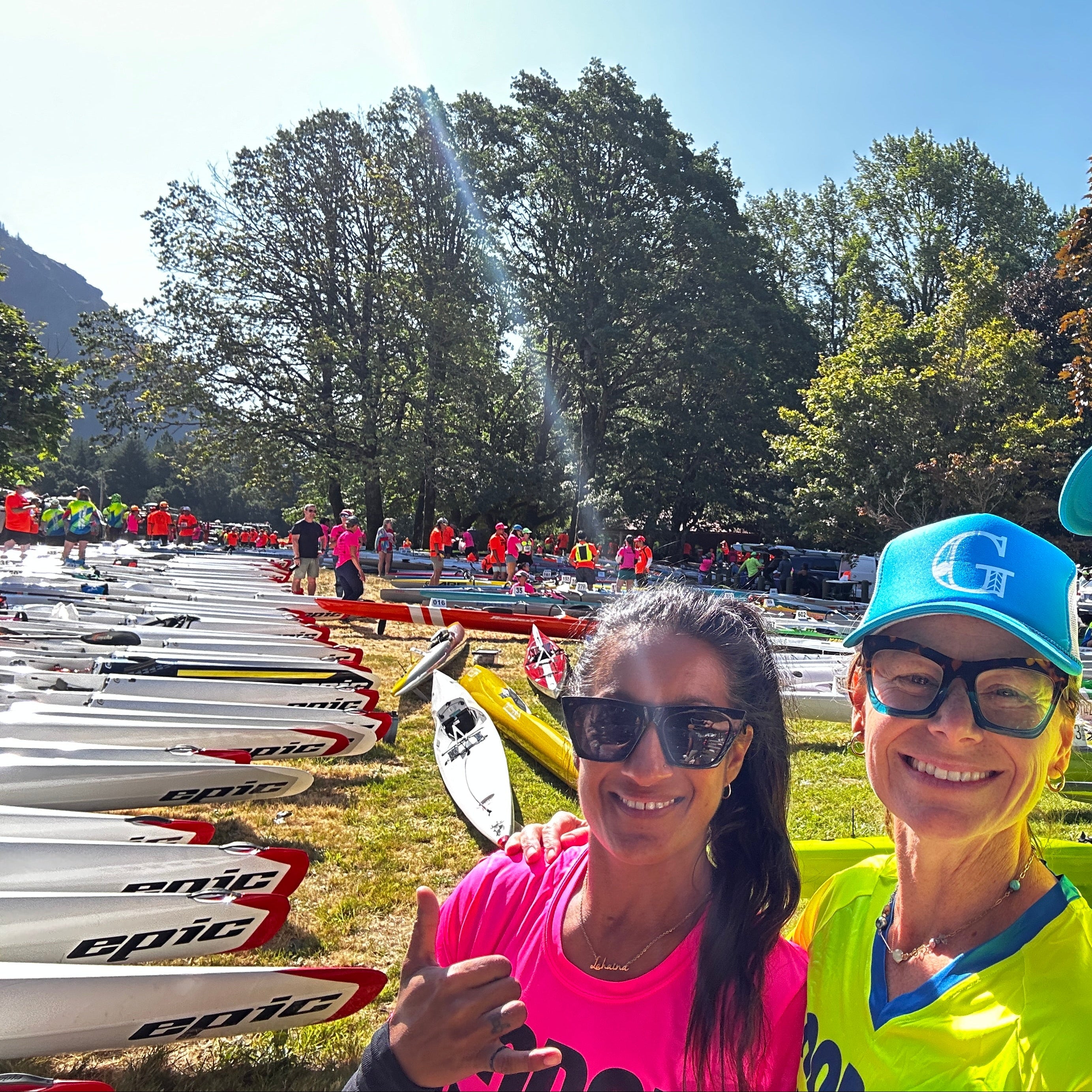 Two people posing for a selfie at a paddling event with kayaks lined up in the background.