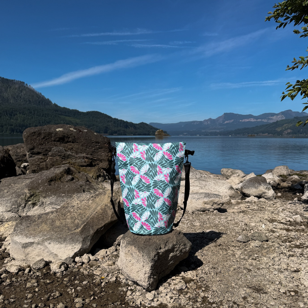 Napili Dry Bag – 20L waterproof wet-dry sack featuring a green palm leaf and pink geometric print, standing unbuckled with shoulder straps on a sunny, rocky lake beach.