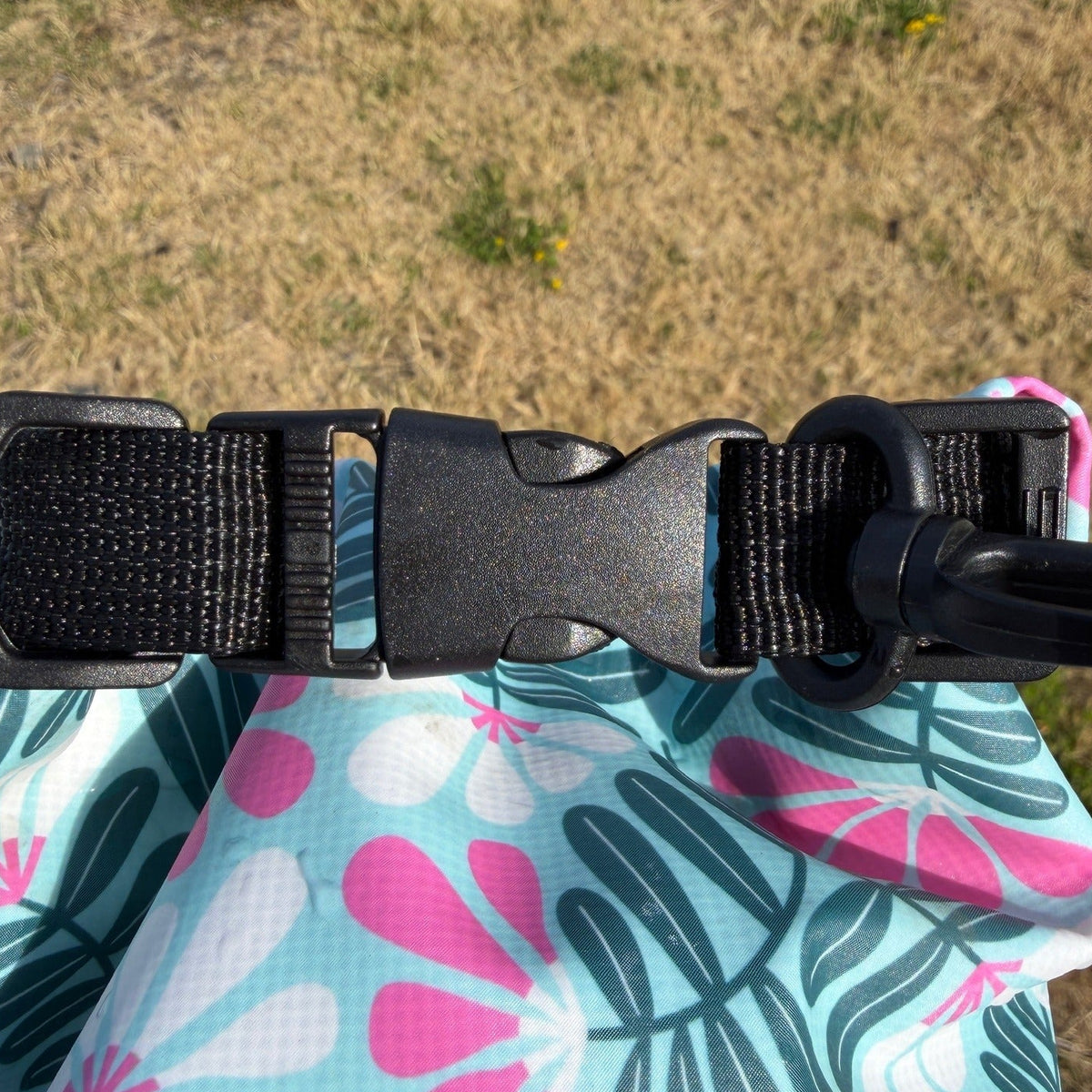 Close-up of a black plastic quick-release buckle attached to a light blue fabric strap with a pink and dark green tropical leaf pattern, set against a dry grassy outdoor background.