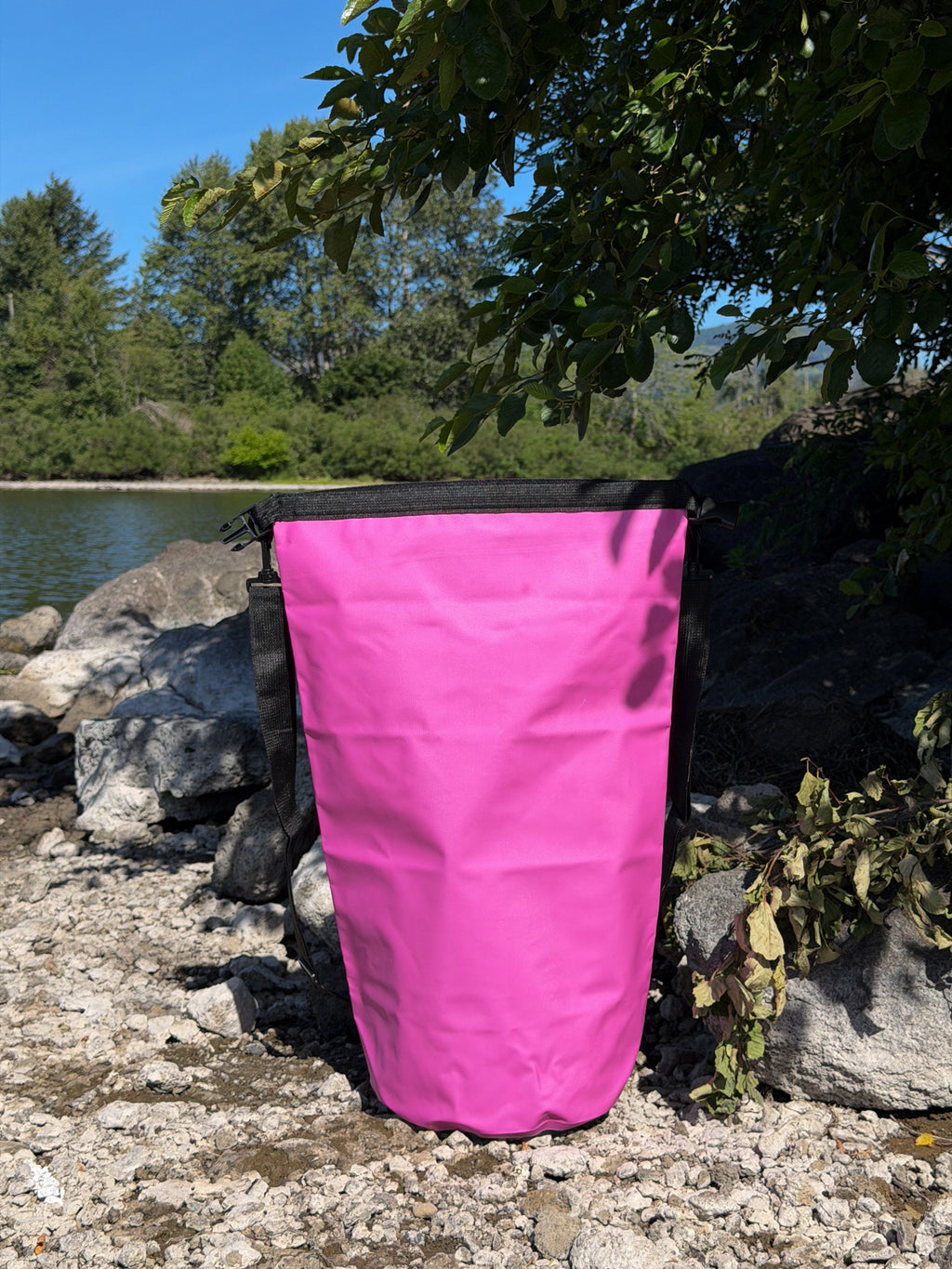 Pismo 20L pink outdoor dry bag with an open flat roll-top standing upright on a gravel and pebble shore under overhanging green tree branches.