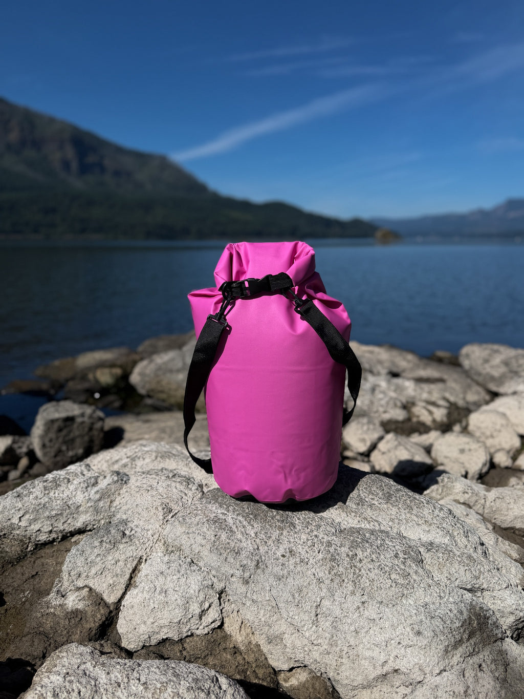 Pismo 20L hot pink waterproof dry sack securely buckled closed, standing on a large gray rock by a scenic lake shoreline under a clear blue sky.