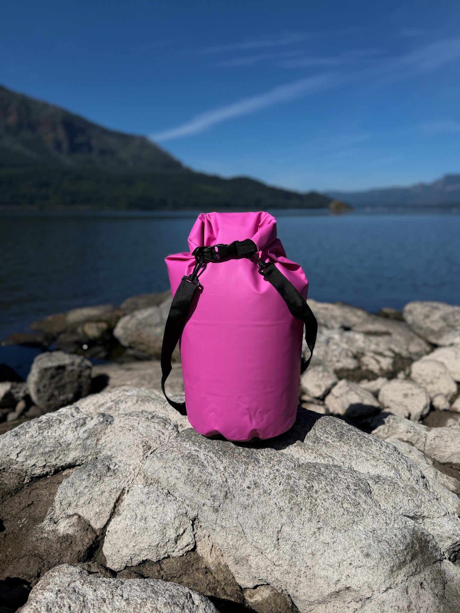Pismo 20L hot pink waterproof dry sack securely buckled closed, standing on a large gray rock by a scenic lake shoreline under a clear blue sky.