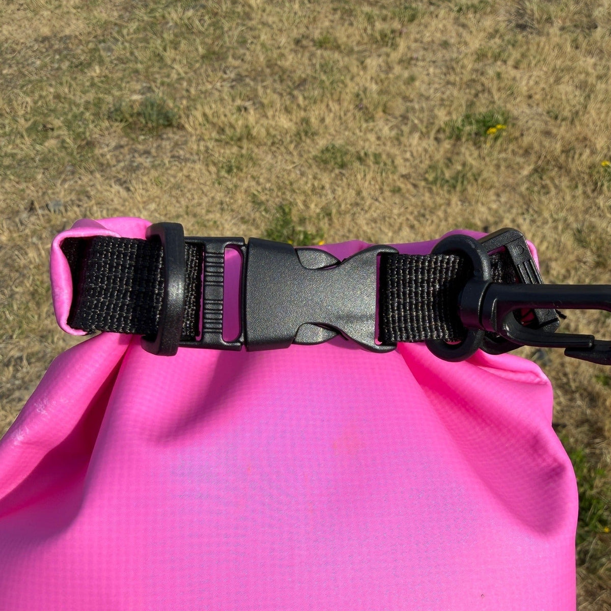 A close-up of a bright pink waterproof bag with a black plastic buckle and black webbing strap, photographed outdoors on a dry grassy field.