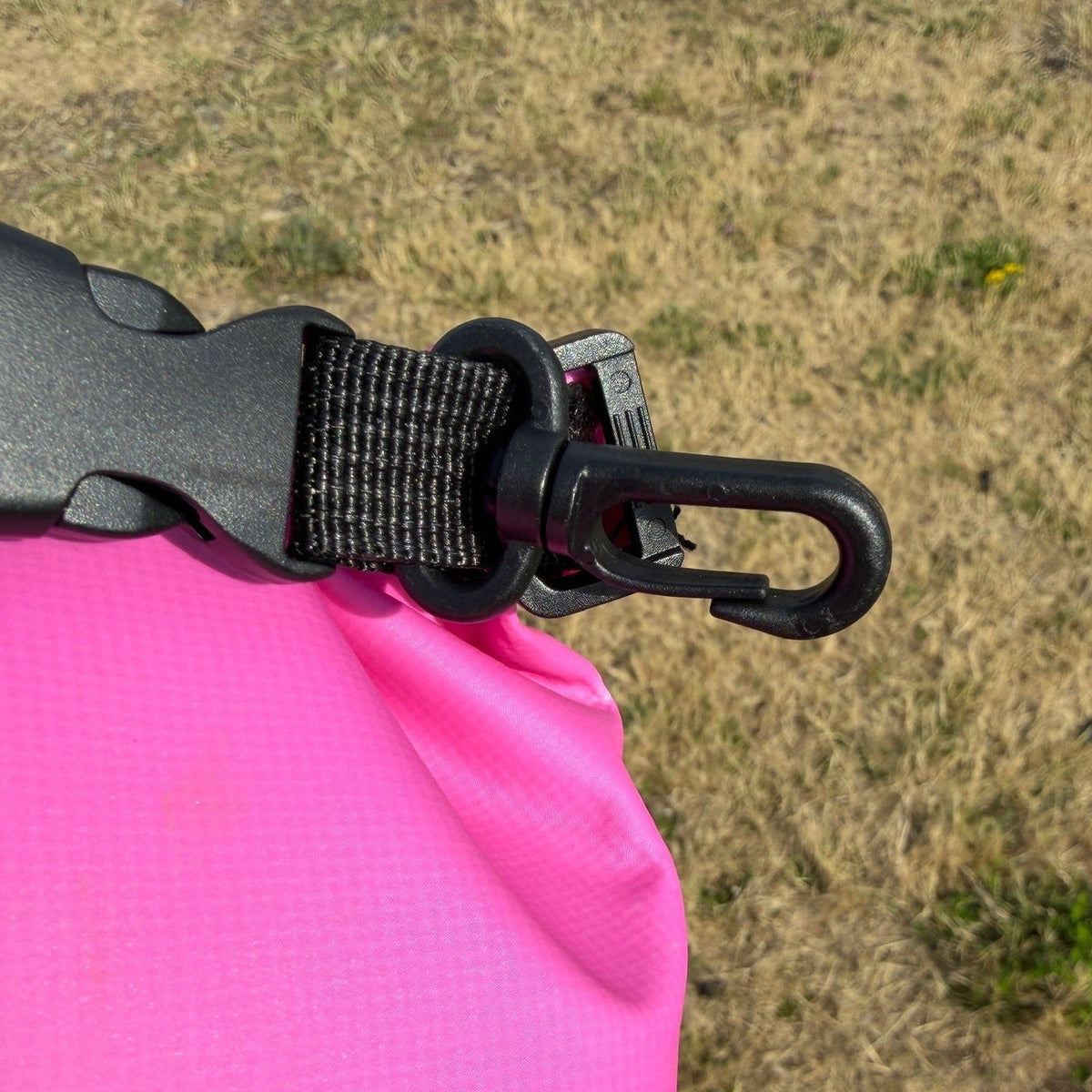 Close-up of a bright pink waterproof bag with a black plastic buckle and plastic clip attachment, photographed outdoors against dry grass.