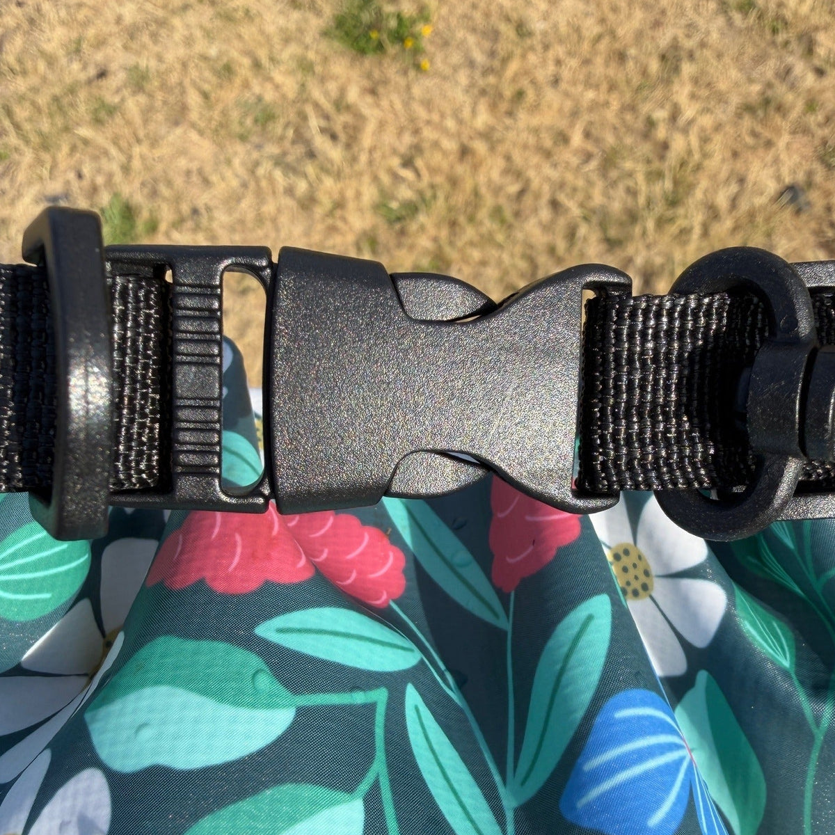 Close-up of a black plastic quick-release buckle attached to a black strap over a dark green floral fabric with blue, green, pink, and white leaf and flower patterns, with dry grass visible in the background.
