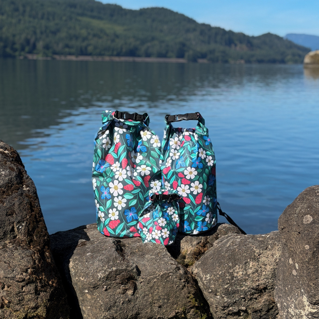 A collection of outdoor wet-dry gear including the Spaulding Dry Bag – 20L floral sack and matching smaller pouches, displayed on large rocks next to a calm lake.