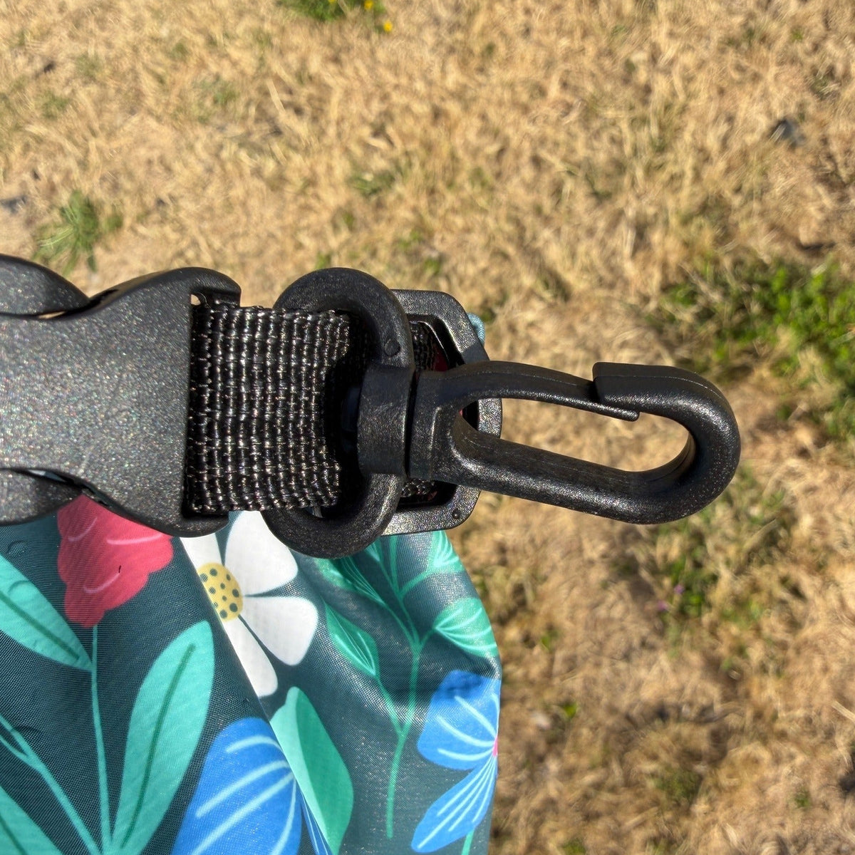Close-up of a black plastic carabiner clip attached to a dark green waterproof bag with a blue, green, and white floral pattern, set against a dry grassy outdoor background.