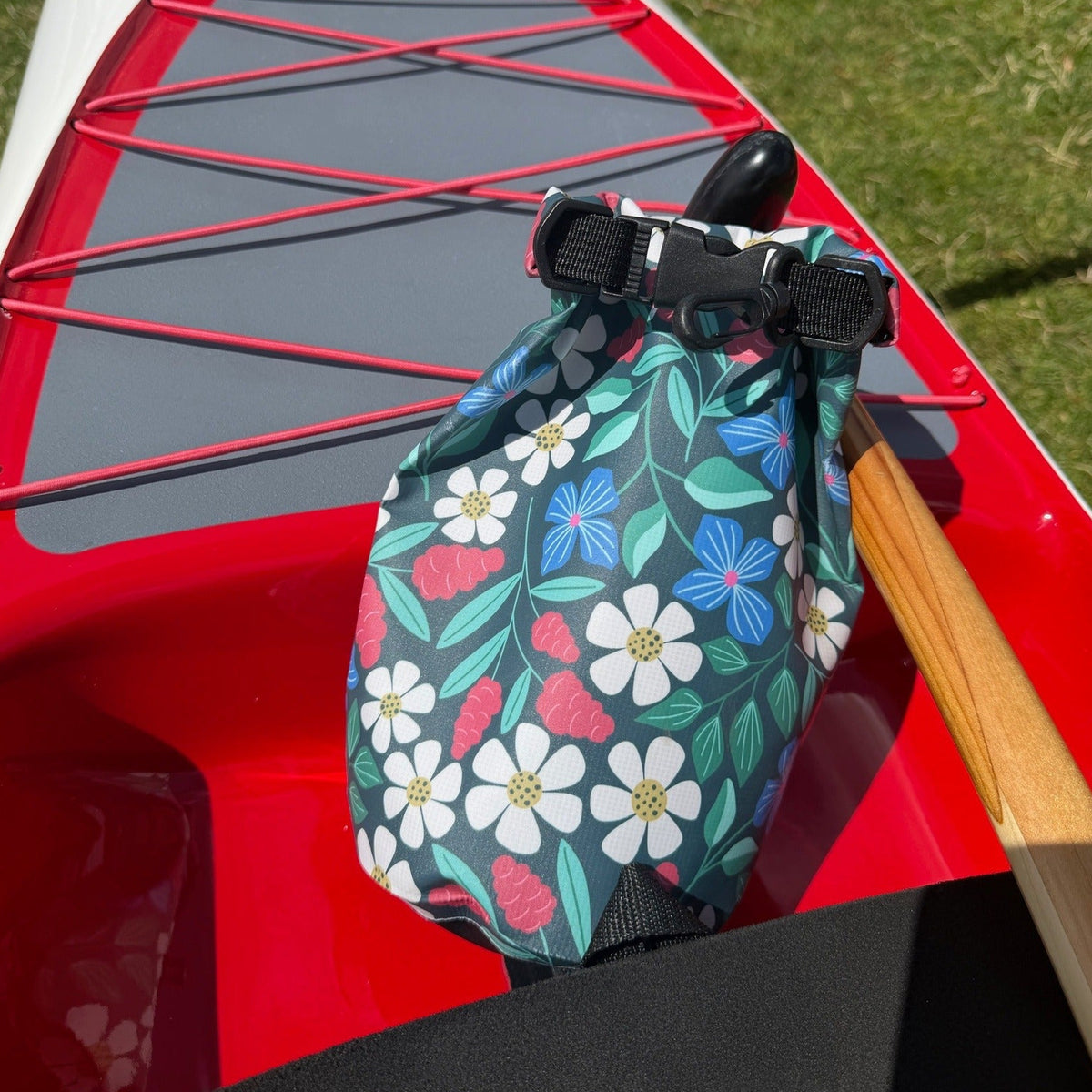 A floral-patterned dry bag with black plastic buckles and straps, resting on the red hull of a canoe outdoors on grass.