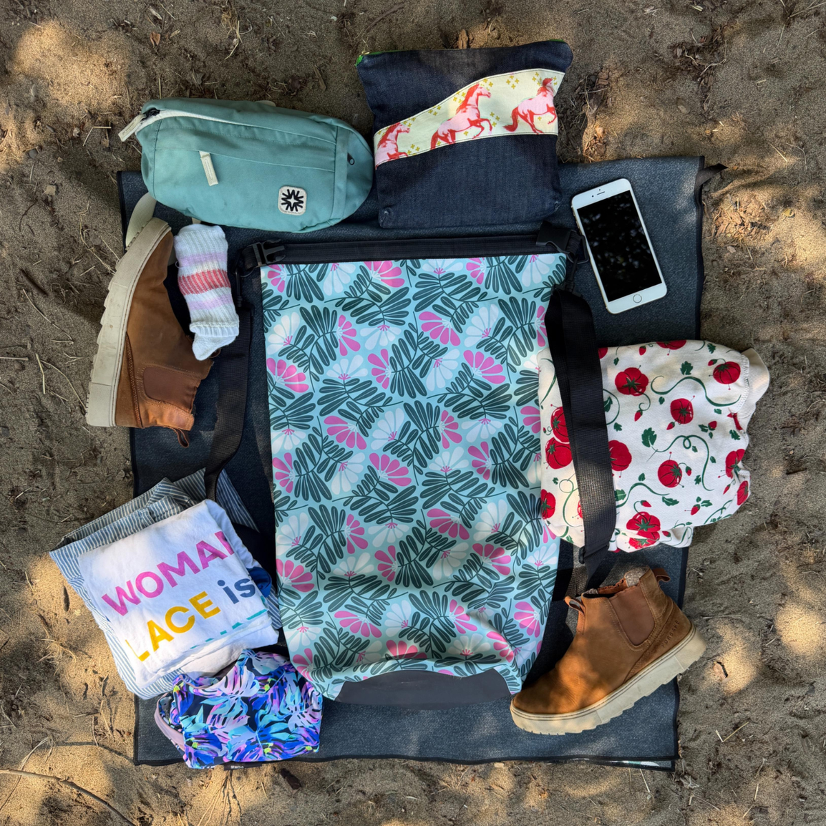 A top-down flat lay of a woman's travel or camping gear set arranged on a black sleeping pad over sandy ground. The set includes a large central reusable tote bag with a green palm leaf pattern, a mint green pouch, a dark denim pouch, a white phone case, a pair of brown boots, a striped sock, a colorful t-shirt, and a red-and-white patterned cloth.