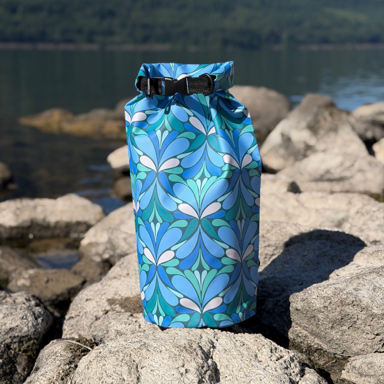 Yuba 20L roll-top dry bag with a vibrant blue and teal pattern sitting on a rocky lake shoreline during a sunny outdoor adventure.