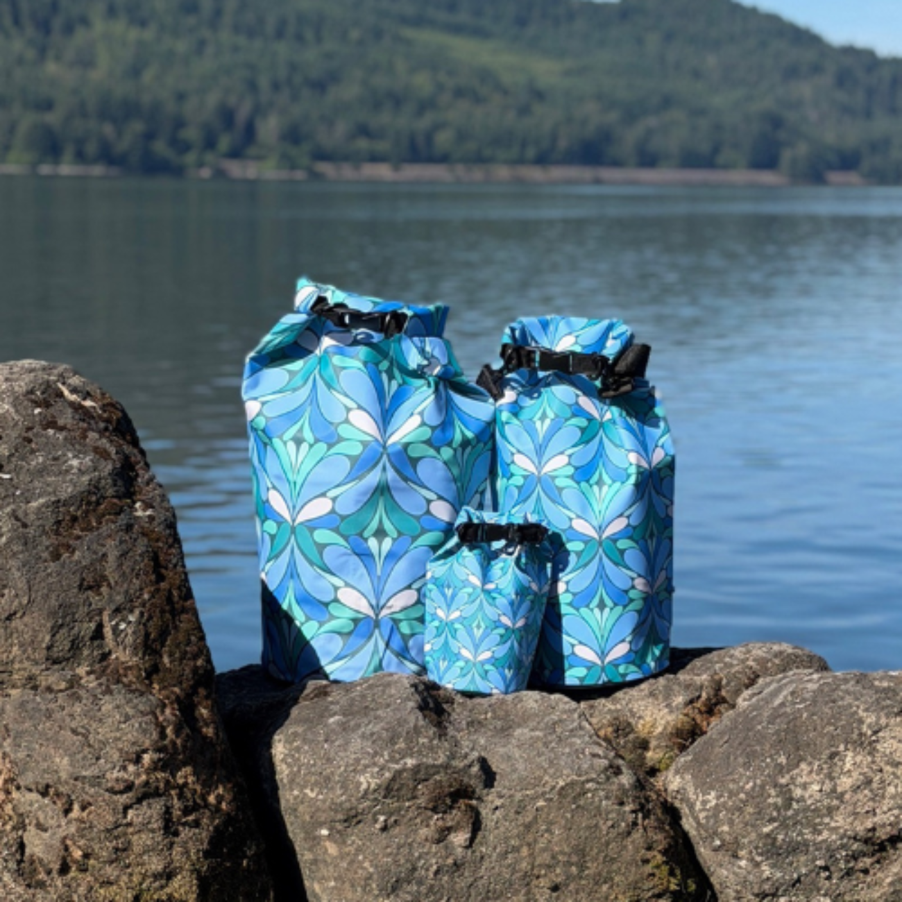 Three different sizes of blue patterned dry bags on rocks by a lake with trees in the background