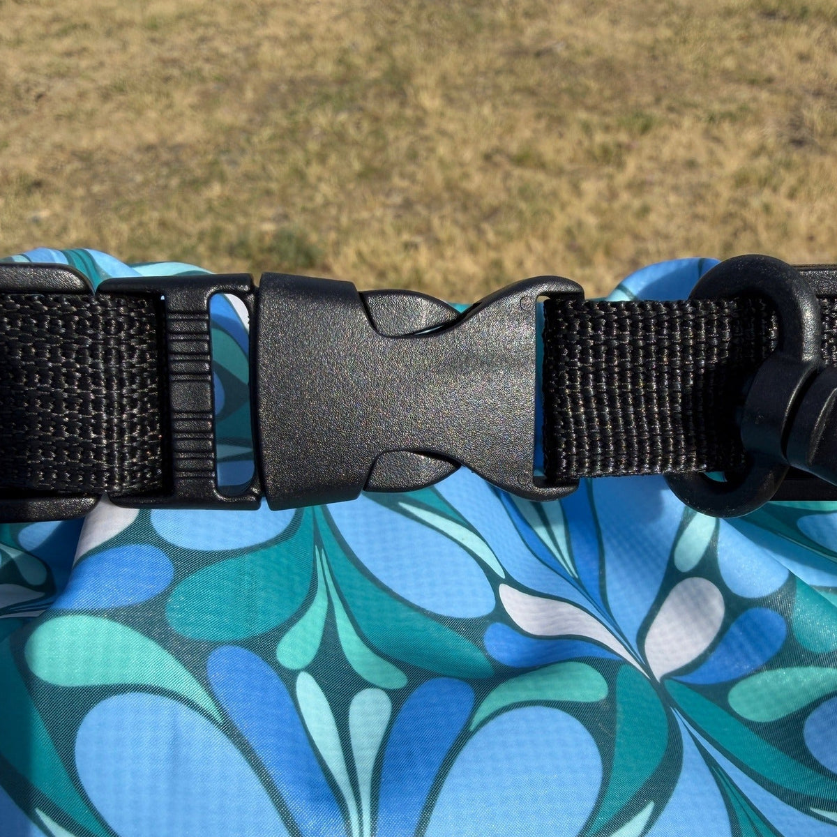 Close-up of a patterned dry bag with a black plastic quick-release buckle and black webbing strap, set against a blurred outdoor grassy background.