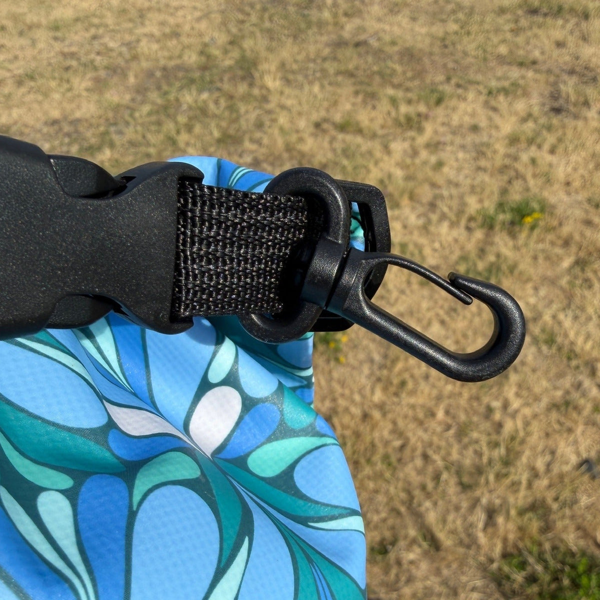 Close-up of a blue and teal geometric-patterned bag with a black plastic buckle and black carabiner clip, photographed outdoors against dry grass.