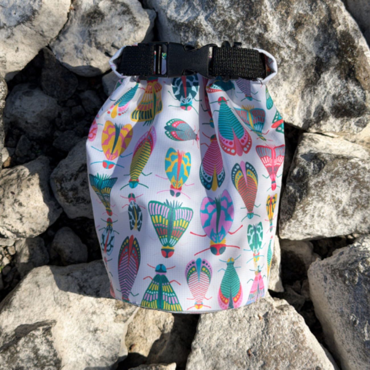 A white dry bag with a bright insect pattern sitting upright on rugged gray rocks under bright sunlight.