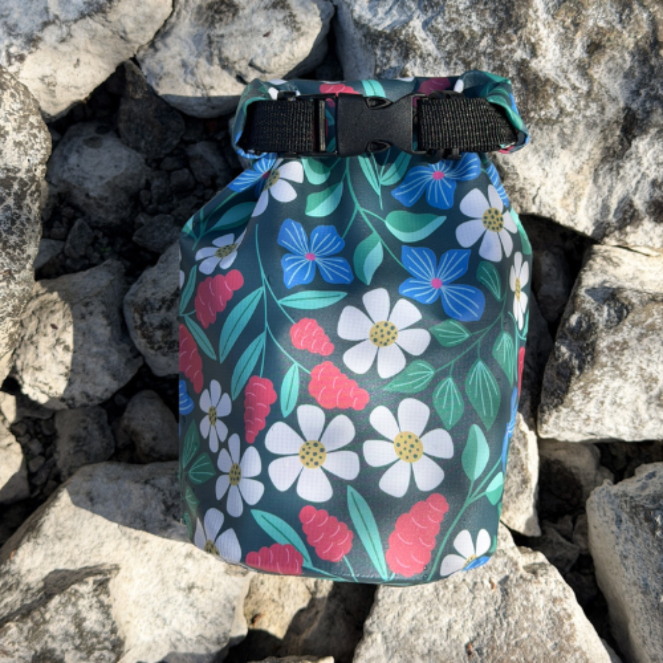 A compact floral print waterproof bag sitting vertically amidst textured gray rocks on an outdoor excursion.