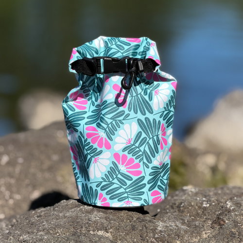 A small waterproof dry bag with a pink and green botanical print sitting on a dark rock by the water.
