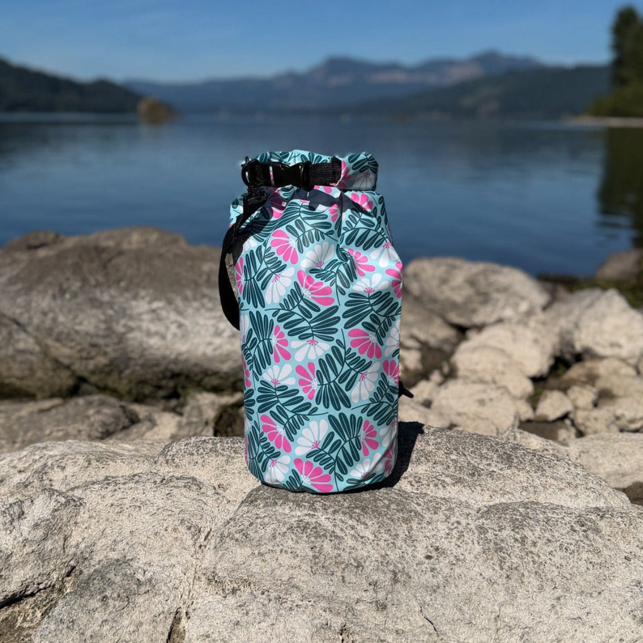 The tropical print Napili 10L roll-top dry bag sitting on sunny grey rocks along a shoreline, with a calm lake and forested mountains under a blue sky in the background.