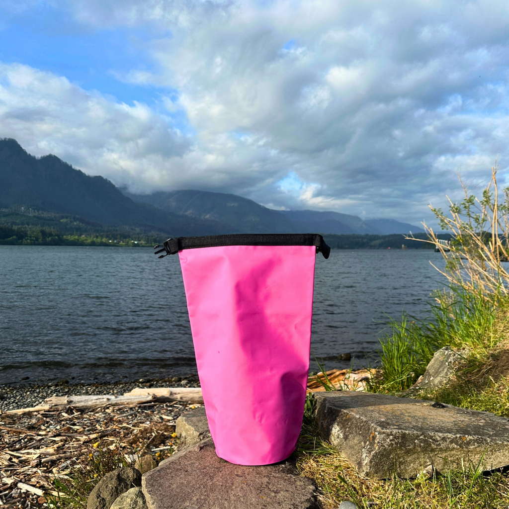 Pink waterproof wet-dry sack shown unbuckled with an open roll-top design, resting on a rocky beach next to a calm lake with cloudy mountain scenery.