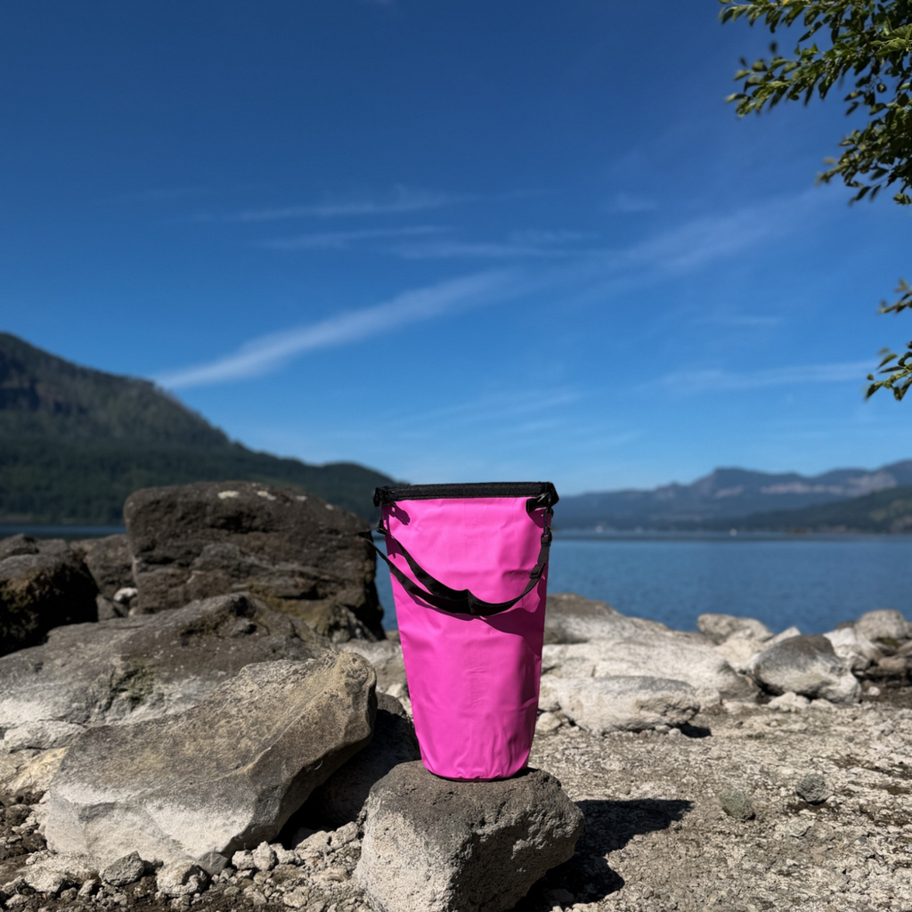 Vibrant pink waterproof outdoor dry bag buckled shut and standing on a rocky lake shoreline with mountains and blue skies in the background.