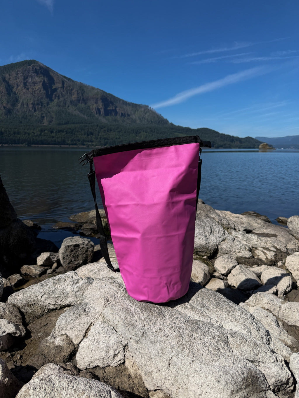 An outdoor lifestyle shot of the pink Pismo 20L Waterproof Dry Bag standing unrolled on a rocky lake shoreline, showcasing the bag's capacity against a scenic background of water and mountains.