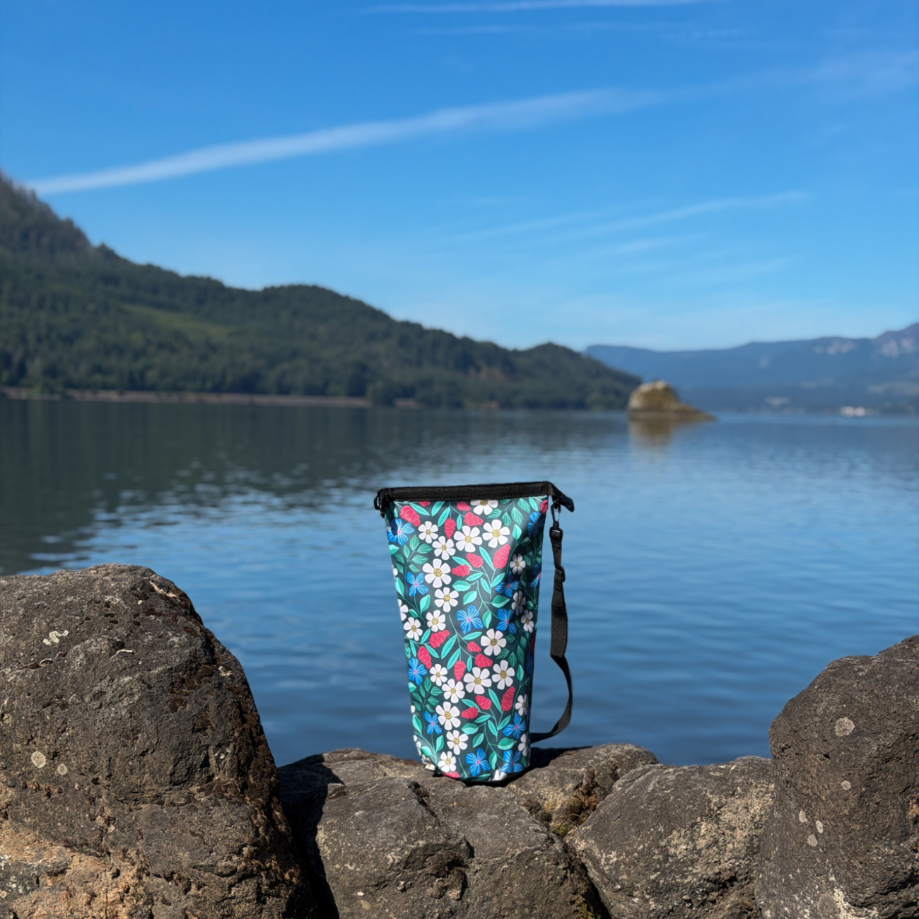 Side view of the Spaulding 10L waterproof dry bag resting on dark stones by the lakeside, showing the slim profile of the bag under a clear blue sky.