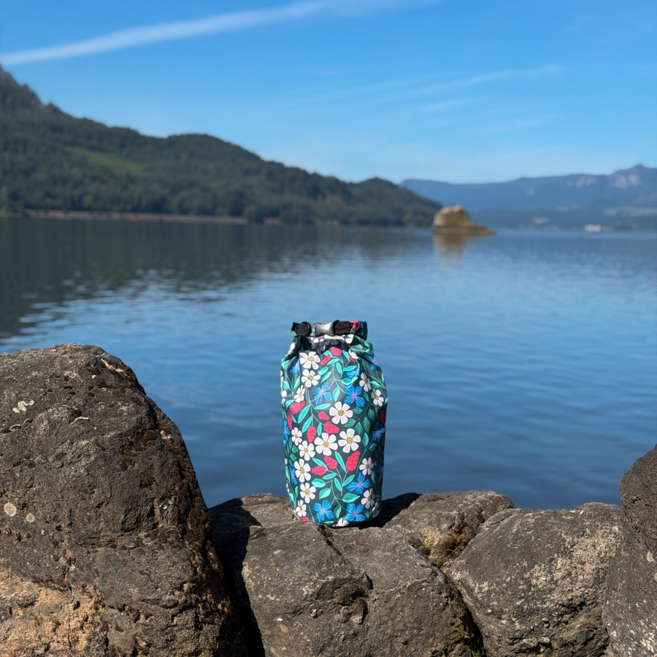 A floral 10L Waterproof Dry Bag resting on lakefront rocks with a scenic view of calm blue water and distant mountains under a clear sky.