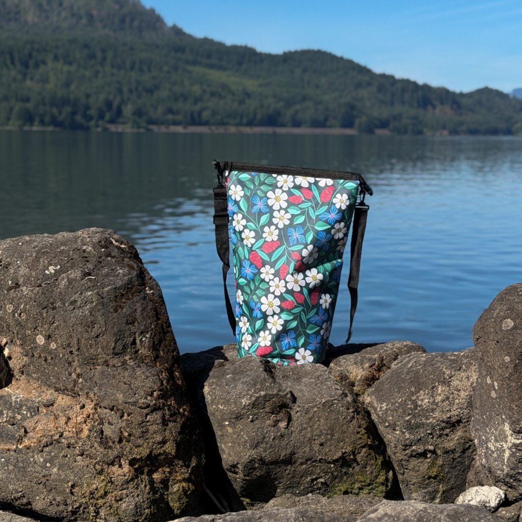 The floral Spaulding 20L Waterproof Dry Bag standing open and unrolled on a rocky beach, showcasing its wide top opening for easy packing. The 20-liter dry sack is nestled between large lakeside boulders under a bright sky.