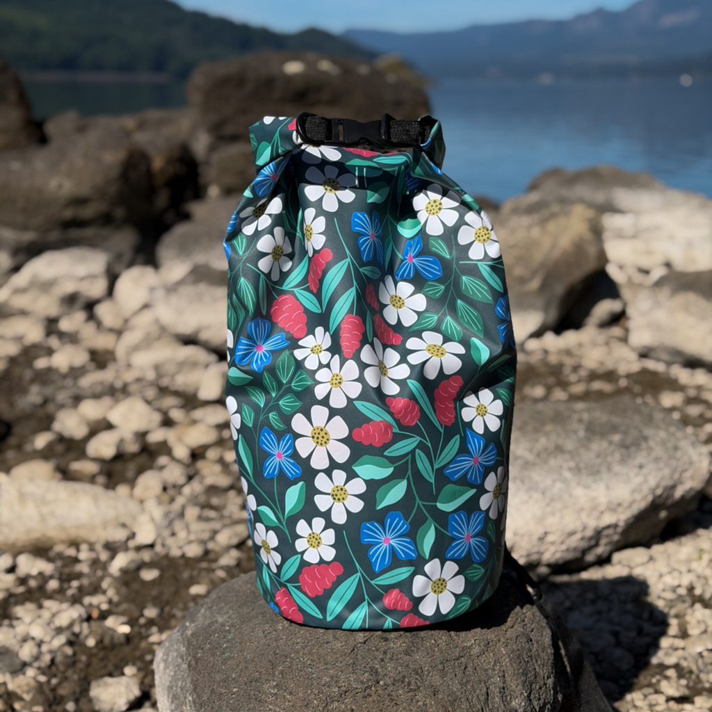 The Spaulding 20L Waterproof Dry Bag standing on a rugged, sun-drenched stone shore next to a calm lake. This durable, leakproof roll-top dry bag keeps camping essentials and electronics safe and dry during outdoor water sports, shown against a mountain lake backdrop.