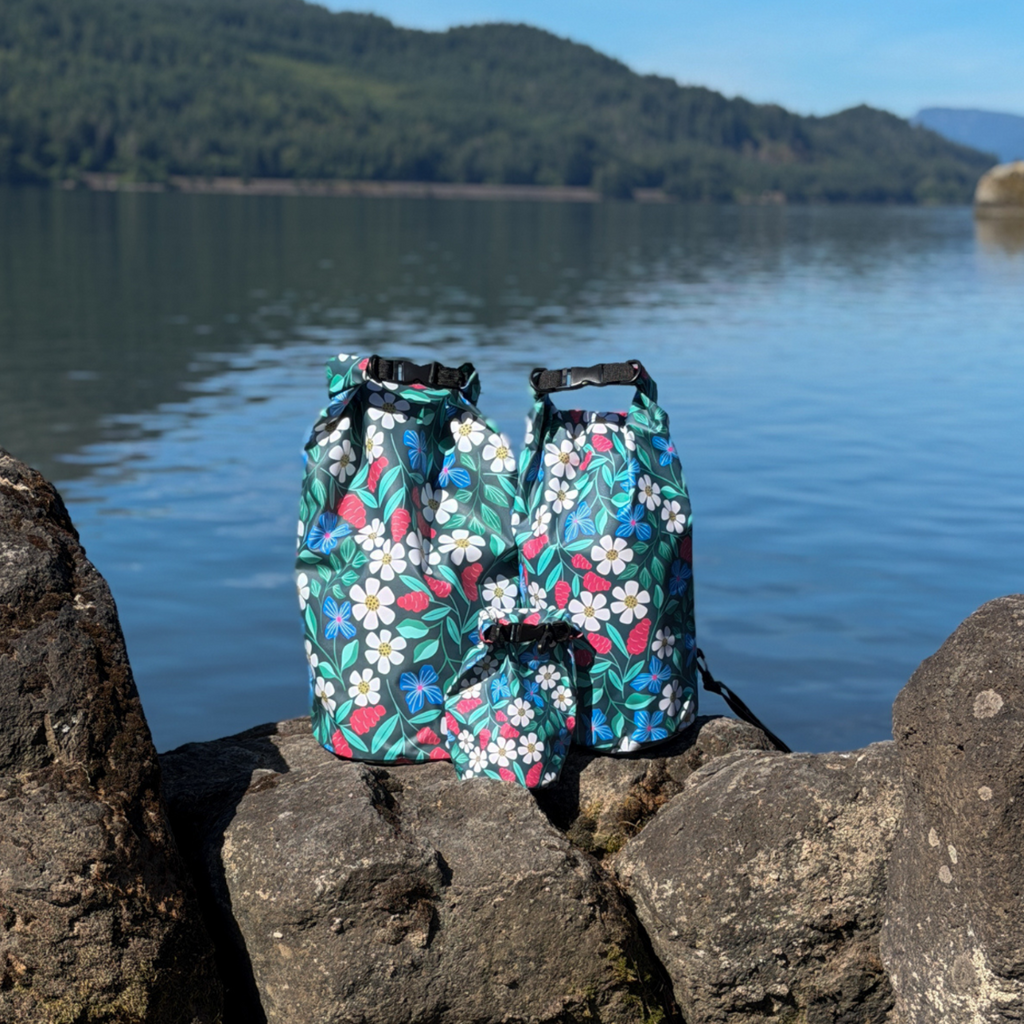Two matching Spaulding 20L Waterproof Dry Bags featuring the floral design, standing upright on dark rocks by a calm lake. Premium waterproof outdoor gear perfect for paddleboarding (SUP), canoeing, and rafting, set against a background of water and distant hills.