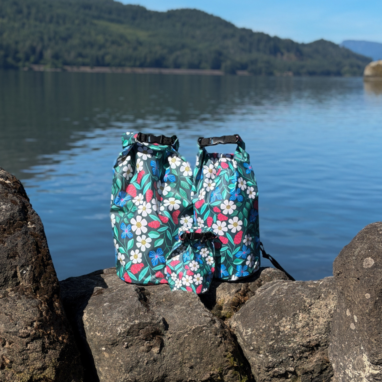 Two matching Spaulding 20L Waterproof Dry Bags featuring the floral design, standing upright on dark rocks by a calm lake. Premium waterproof outdoor gear perfect for paddleboarding (SUP), canoeing, and rafting, set against a background of water and distant hills.