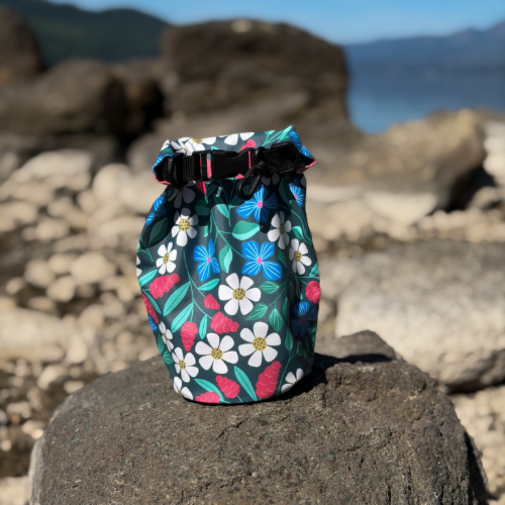 A daisy pattern waterproof dry sack placed on a dark boulder along a scenic rocky shoreline with lake and hills in the background.