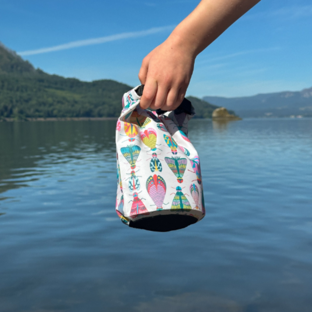 A person's hand holding a colorful bug-themed waterproof dry bag over clear lake water with mountains and blue sky in the background.