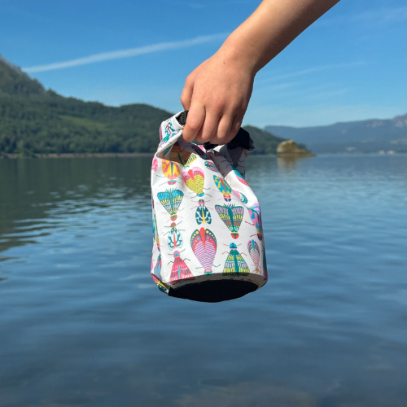 A person's hand holding a colorful bug-themed waterproof dry bag over clear lake water with mountains and blue sky in the background.