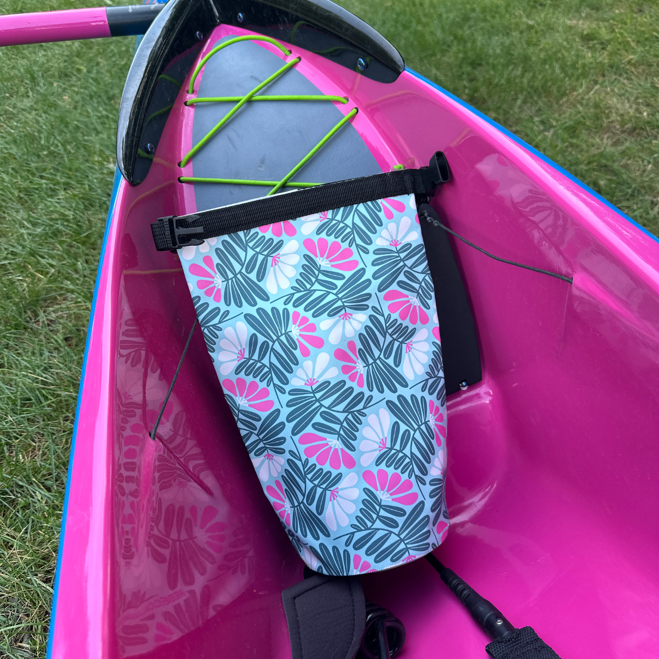A pink and green tropical pattern dry bag tucked securely inside the cockpit of a bright pink kayak on the grass.