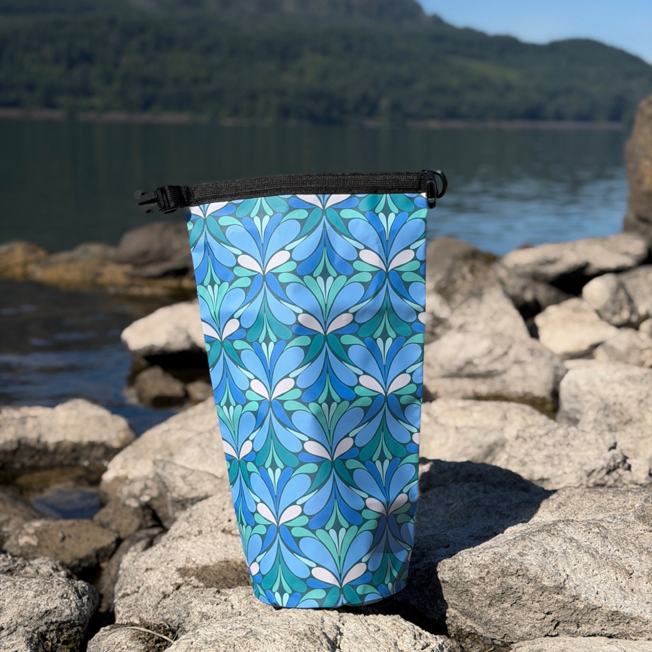 An outdoor lifestyle photo of the Yuba 10L Waterproof Dry Bag standing unrolled and open on a rocky beach, showcasing its pattern against a scenic background of water and distant green mountains.