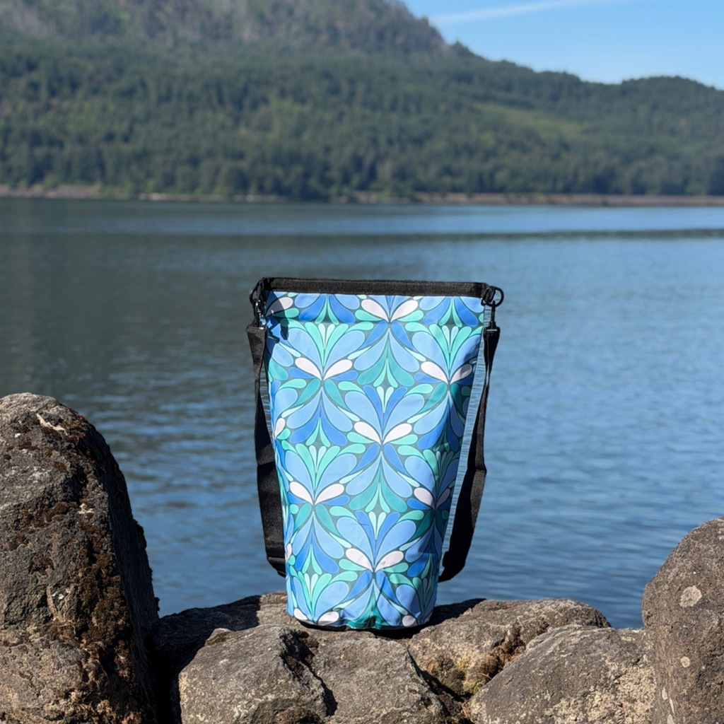 Side view of the patterned Yuba waterproof dry bag showing the attached black straps, placed on a rocky retaining wall next to a lake under a clear blue sky.