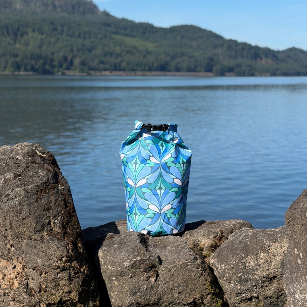 The blue patterned Yuba 20L dry bag standing on dark rocks along a sunny shoreline, with a calm lake and forested hills in the background.